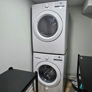 A stacked washer and dryer unit in a laundry room with a black table in front. - Carekaters Haven | Cozy 1BR • Wi-Fi • Free Parking (Saskatoon)