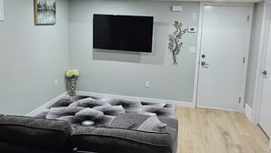 Ein Wohnzimmer mit grauem Sofa, einem an der Wand montierten Flachbildfernseher, einer Tür und einer kleinen Zimmerpflanze.