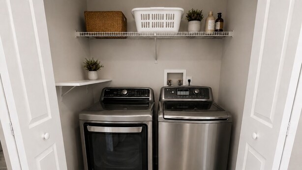 A compact laundry area with two stainless steel washers and dryers, a white shelf above with baskets and toiletries, and a small potted plant.