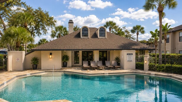 A swimming pool with a stone border, a covered seating area with chairs, and a building with a tiled roof.
