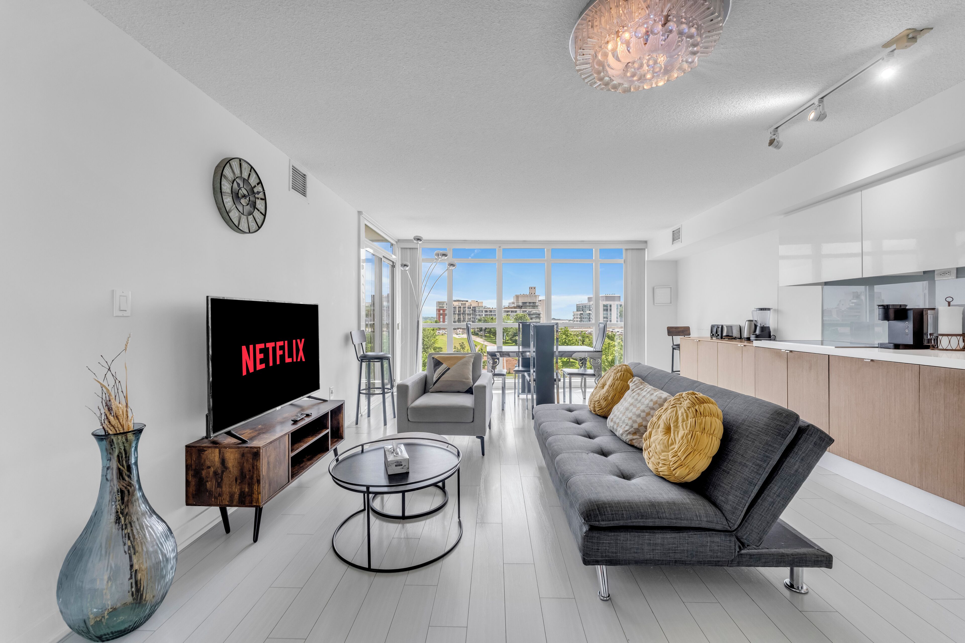 A modern living room with a flat-screen TV, a sofa, a coffee table, and a view of the city through large windows.