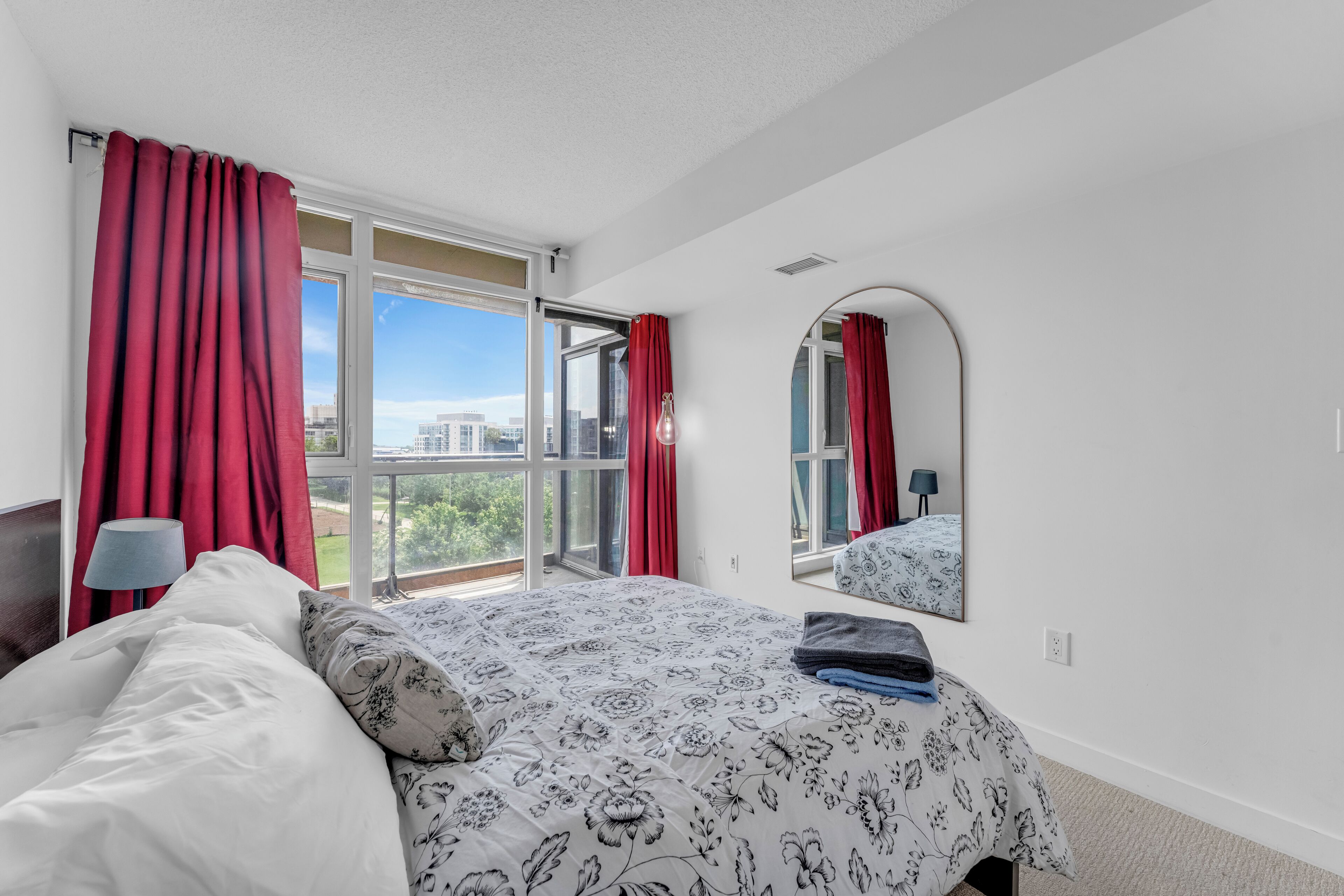 A bedroom with a large bed, a mirror, and a view of buildings through the window.