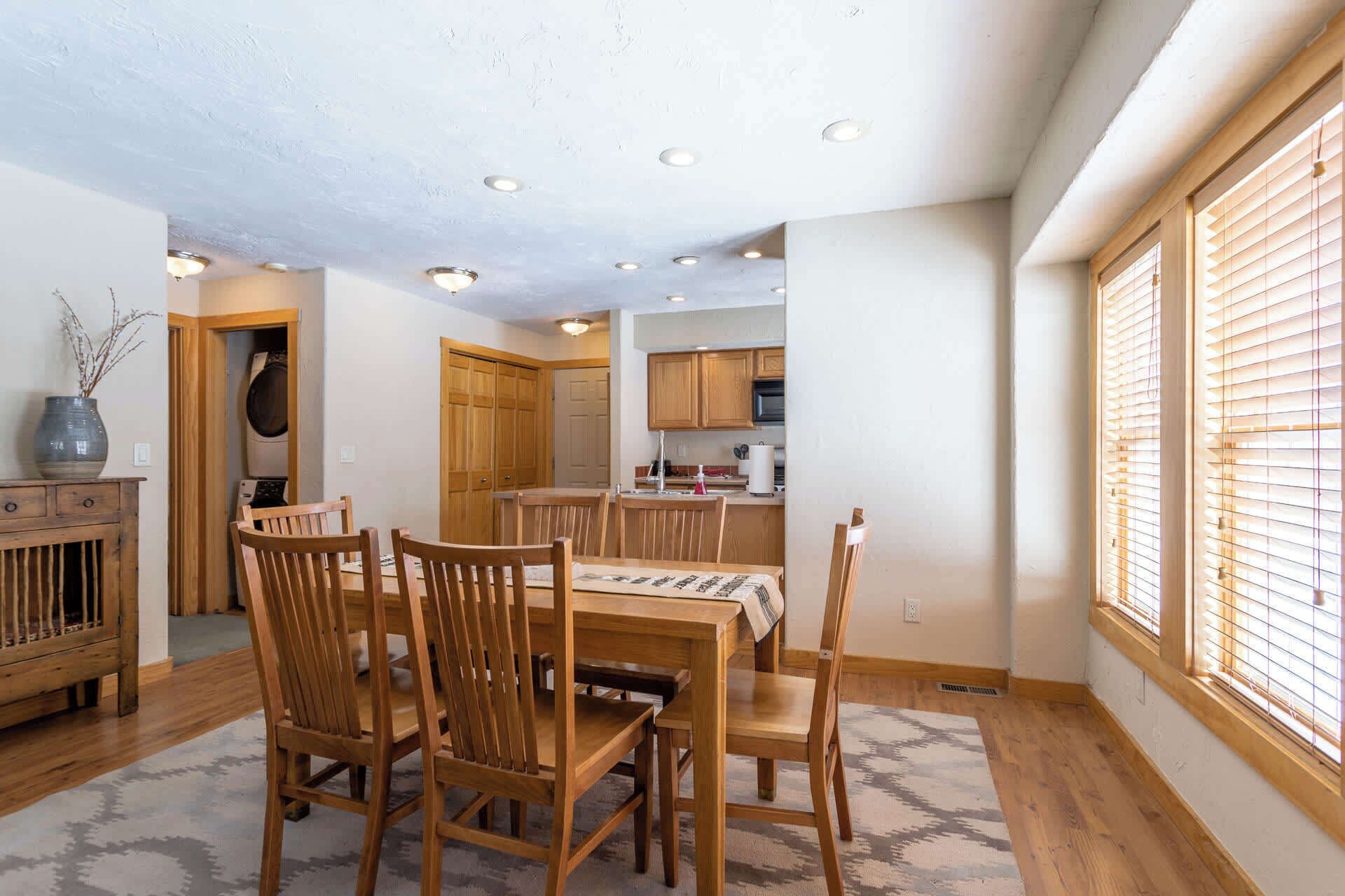 A dining area with wooden chairs and a table, a kitchen with wooden cabinets, and a window with blinds.