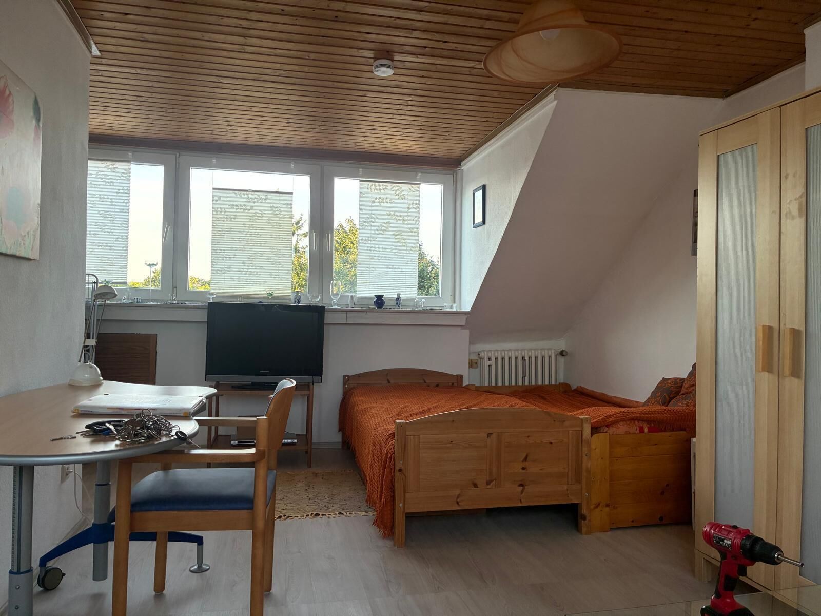 Ein Schlafzimmer mit schrägen Wänden, einem Bett, einem Schreibtisch mit Stuhl, einem Fernseher und einem Fenster mit Jalousien.