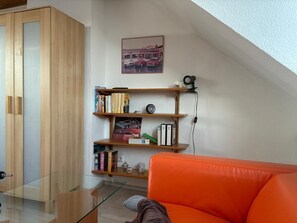 A room with a glass table, an orange sofa, wooden shelves with books, a picture frame, and a lamp. - TRADE FAIR DÜSSELDORF APARTMENT (Duisburg)