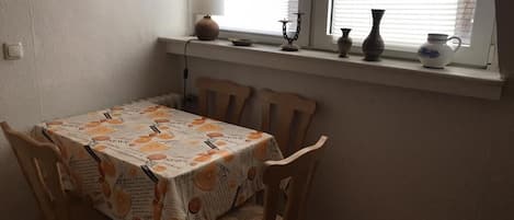 Un comedor con una mesa dispuesta para cuatro personas, una ventana con persianas y un estante con objetos decorativos.