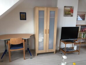 A room with a wooden cabinet, a desk with a chair, a television on a stand, and framed pictures on the wall. - TRADE FAIR DÜSSELDORF APARTMENT (Duisburg)