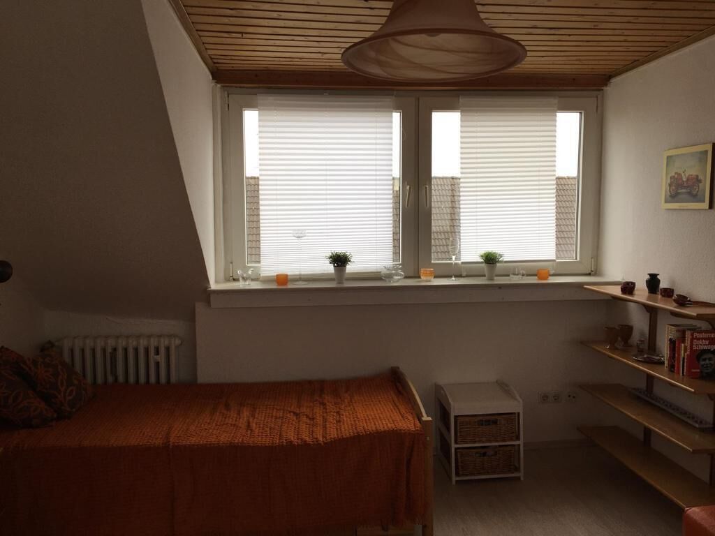 A bedroom with a sloped ceiling, a window with blinds, a bed with a blanket, a radiator, and a shelf with books and decorative items.