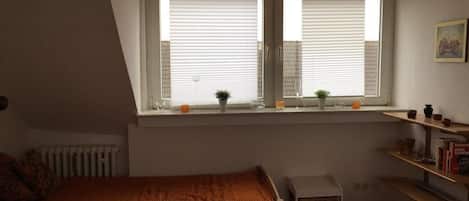 A bedroom with a sloped ceiling, a window with blinds, a bed with a blanket, a radiator, and a shelf with books and decorative items.