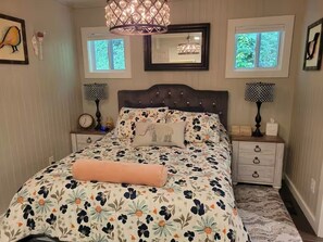 A bedroom with a large bed, two bedside tables, a nightstand, a mirror, and a window with a view of greenery. - Charming cottage in the mountains of NE Georgia, five minutes from Clayton, GA. (Clayton)