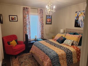 A bedroom with a bed, a red armchair, a chandelier, and a window with curtains. - Charming 2 bedroom, 2 bath cottage in the mountains of Clayton, Georgia. (Clayton)