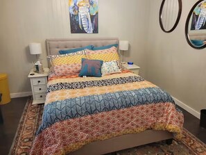 Una cama con colcha estampada y varias almohadas, una mesita de noche con lámpara, y un espejo montado en la pared.