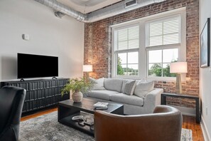 Een moderne woonkamer met een bakstenen muur, een flatscreen televisie, een grijze bank en twee vloerlampen.
