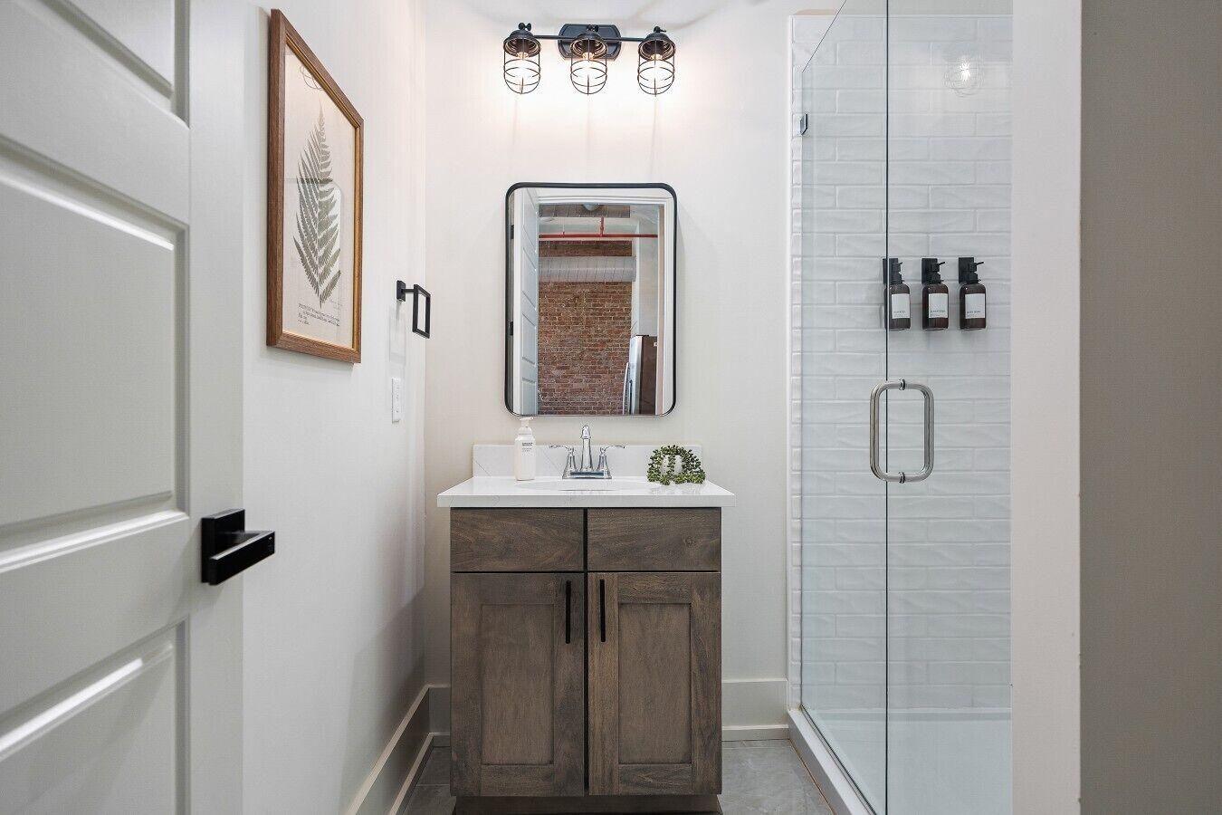 Un baño con tocador blanco, un espejo, ducha con puerta de cristal y una obra de arte con hojas enmarcada en la pared.