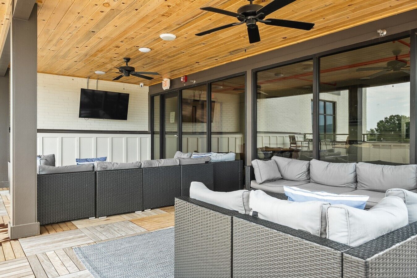 An outdoor seating area with a ceiling fan, a television mounted on the wall, and a view of the outside through large windows.