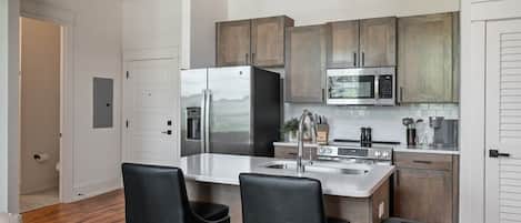 A modern kitchen with stainless steel appliances, wooden cabinets, and a bar area with two black chairs.
