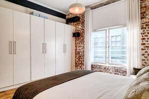 A bedroom with a large bed, a wardrobe, a brick wall, and a window with curtains. - Central City 2bed With Mountain Views Skywood Loft (Cape Town)