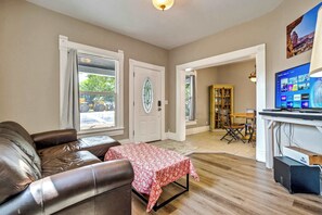 A living room with a brown leather couch, a red and white patterned table, and a television displaying a home screen. - Walnut Porch! Cozy Historic Home Near OCC (Colorado Springs)