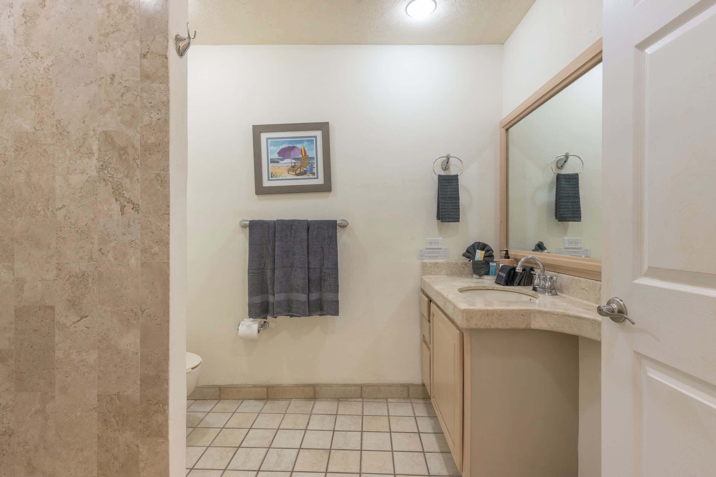 Un baño con inodoro, lavamanos y espejo. Hay un perchero con toallas y un cuadro en la pared.