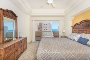 Ein Schlafzimmer mit einem großen Bett, einer hölzernen Kommode, einem Deckenventilator und Meerblick durch eine Balkontür.