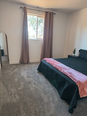 A bedroom with a bed, a window with curtains, a bedside table with a plant, and a carpeted floor.