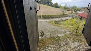 A view from a window overlooking a cobblestone courtyard with a paved path, greenery, and a distant road. - Schist Houses - Casa da Capela (2-Bedroom House) (Bragança)