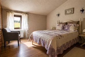 Ein Schlafzimmer mit einem Bett, einem Sessel, einem Fenster mit Vorhängen, einer Wandlampe und einem gerahmten Bild.