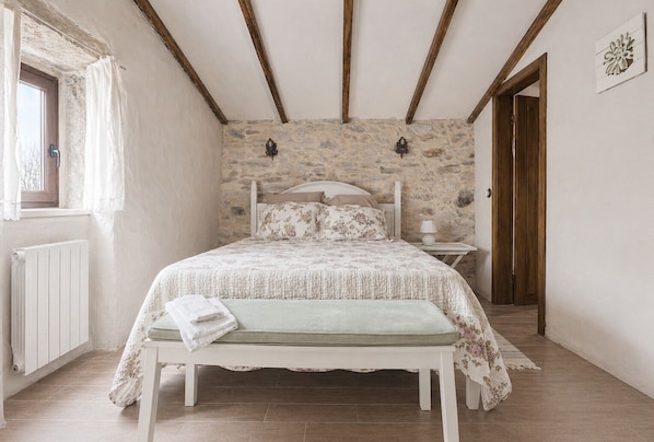 A bedroom with a stone wall, a bed with floral bedding, a bench, a bedside table with a lamp, and a window with curtains.