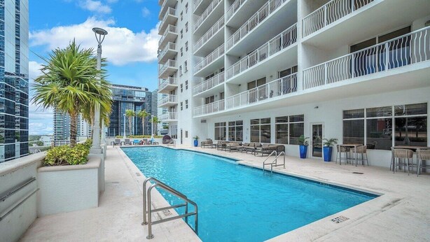 Piscina com escada de metal, rodeada por espreguiçadeiras e mesas, com vista para edifícios altos ao fundo.