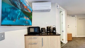 Ein Zimmer mit flachbildfernseher an der Wand, einem Holzkasten mit Schubladen, einer Kaffeemaschine, einem Wasserkocher und einer Klimaanlage.