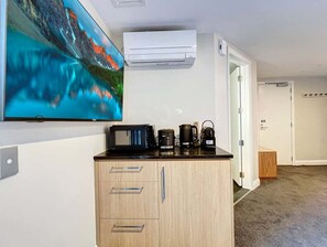 A room with a flat-screen TV mounted on the wall, a wooden cabinet with drawers, a coffee maker, a kettle, and an air conditioning unit. - Above the Lake | Studio Escape (Queenstown)