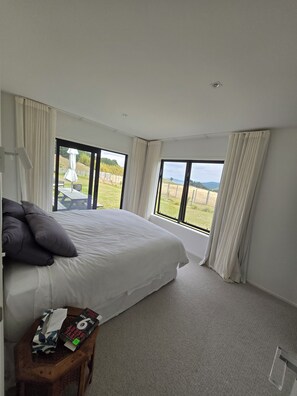Ein Schlafzimmer mit einem großen Bett, einem Holznachttisch und Blick auf eine grüne Landschaft durch große Fenster.