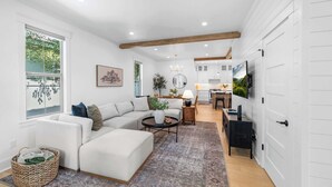 A modern living room with a sectional sofa, a coffee table, and a rug. There is a dining area with a table and chairs, and a kitchen with white cabinets and a large TV mounted on the wall. - Northern Charm with 3BDR & Game Room (Kalispell)