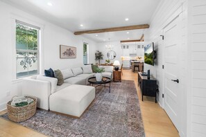 Ein modernes Wohnzimmer mit Ecksofa, Couchtisch und Teppich. Es gibt einen Essbereich mit Tisch und Stühlen sowie eine Küche mit weißen Schränken und einem großen Wand-Fernseher.