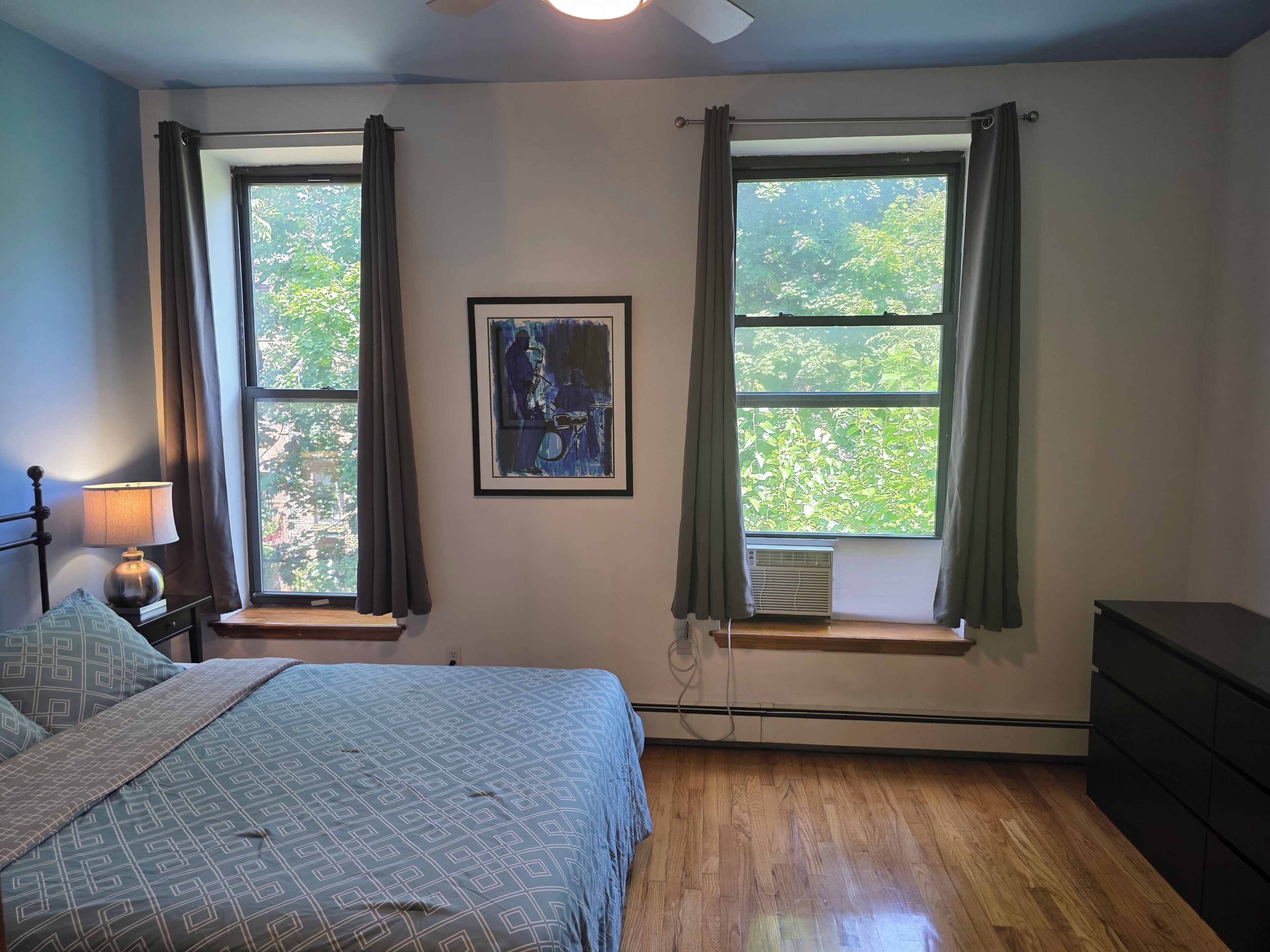 A bedroom with a bed, two windows with curtains, a wall-mounted air conditioner, and a framed picture.