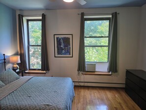 Ein Schlafzimmer mit einem Bett, zwei Fenstern mit Vorhängen, einer Wand-Klimaanlage und einem gerahmten Bild.
