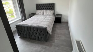 Un dormitorio con cabecera capitonada, una cama grande, dos mesitas de noche, una ventana con cortinas y un radiador.