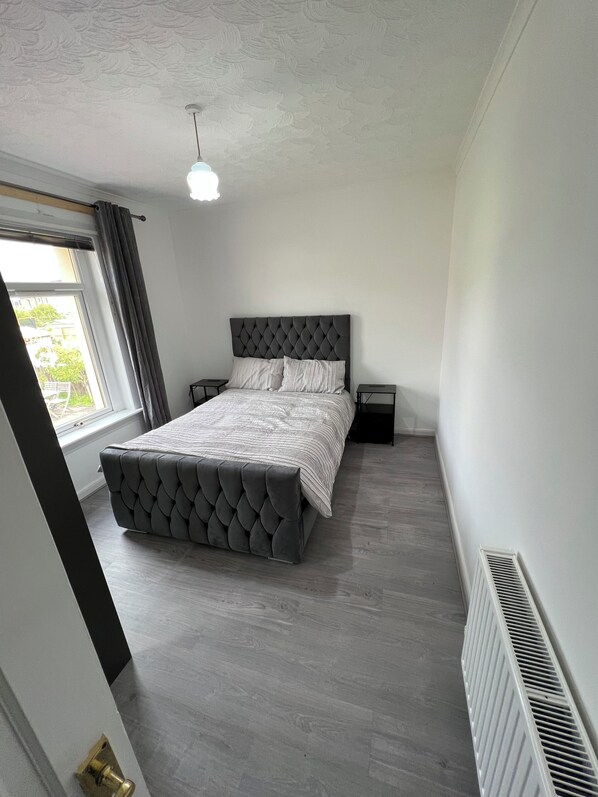 A bedroom with a tufted headboard, a large bed, two bedside tables, a window with curtains, and a radiator.