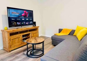 A living room with a flat-screen TV displaying a movie, a grey sofa with yellow cushions, a wooden entertainment unit, and a round stool. - Fast WiFi -3BR2BA Private Yard Work Crew Friendly (Willis)