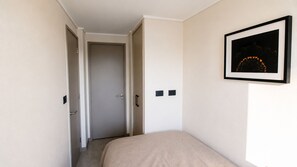 A narrow hallway leading to a door, a bed with a beige cover, and a framed picture on the wall. - Apartment, 2BR/2BA, AC and Heating, parking y GYM. (Ñuñoa)