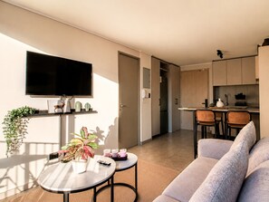Un saló modern amb un sofà, una taula de dinar amb caires, un televisor de pantalla plana fixat a la paret i una cuina amb armaris i taulell.