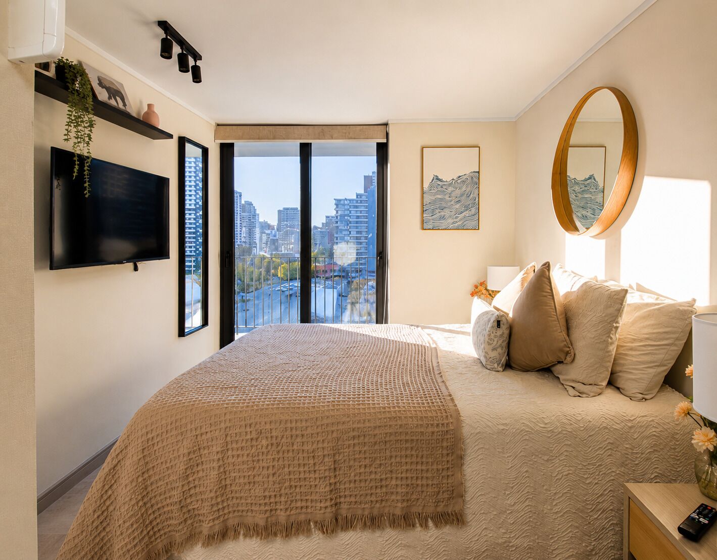 A bedroom with a large bed, a city view, a mounted TV, and a round mirror.