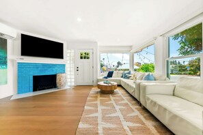 A modern living room with a fireplace, a flat-screen TV, a sofa, and a large window offering a view of the outdoors. - The Tourmaline Beach House - Walk to Surf & Sand (San Diego)