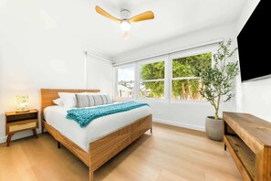 A bedroom with a bed, a ceiling fan, a nightstand, a bedside table, a window with a view of greenery, and a potted plant. - The Tourmaline Beach House - Walk to Surf & Sand (San Diego)
