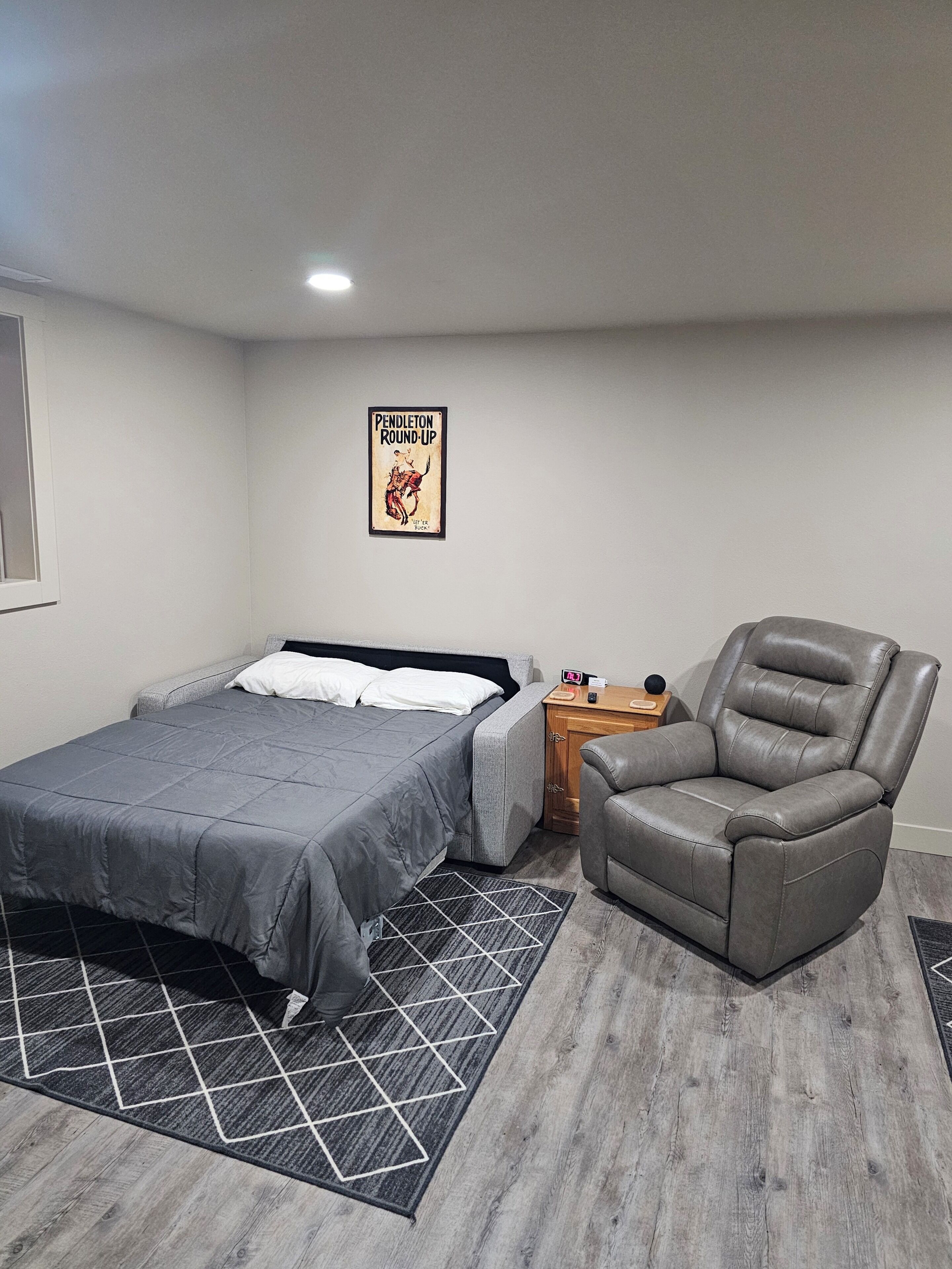 Un dormitorio con una cama, una silla gris, una mesita de noche y un cartel en la pared.