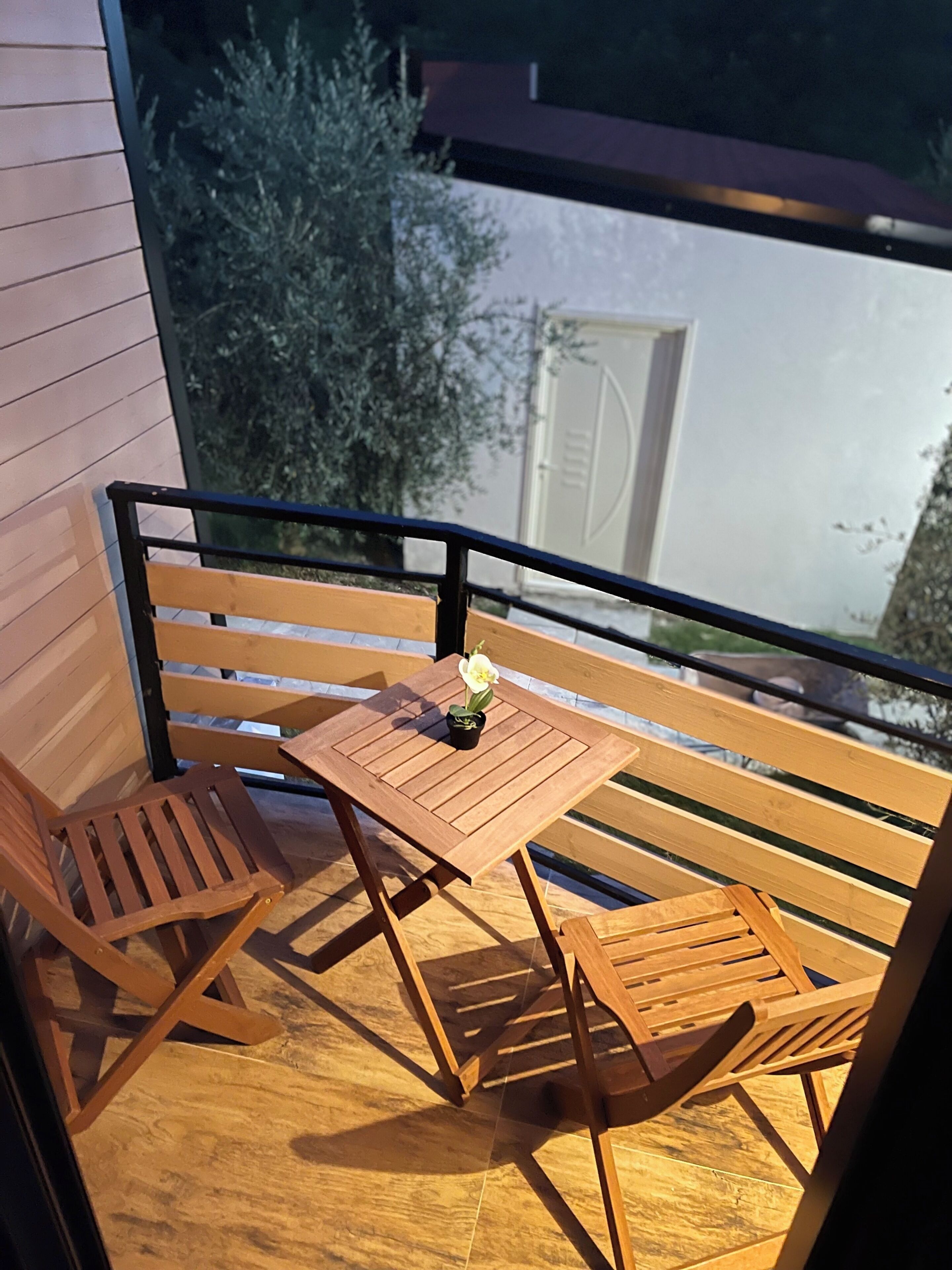 A wooden balcony with a table and two chairs, a small potted plant, and a view of a building with a white door.