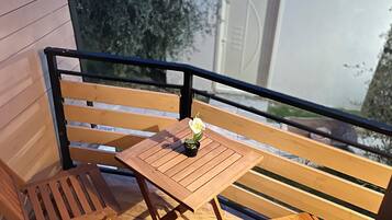 A wooden balcony with a table and two chairs, a small potted plant, and a view of a building with a white door.