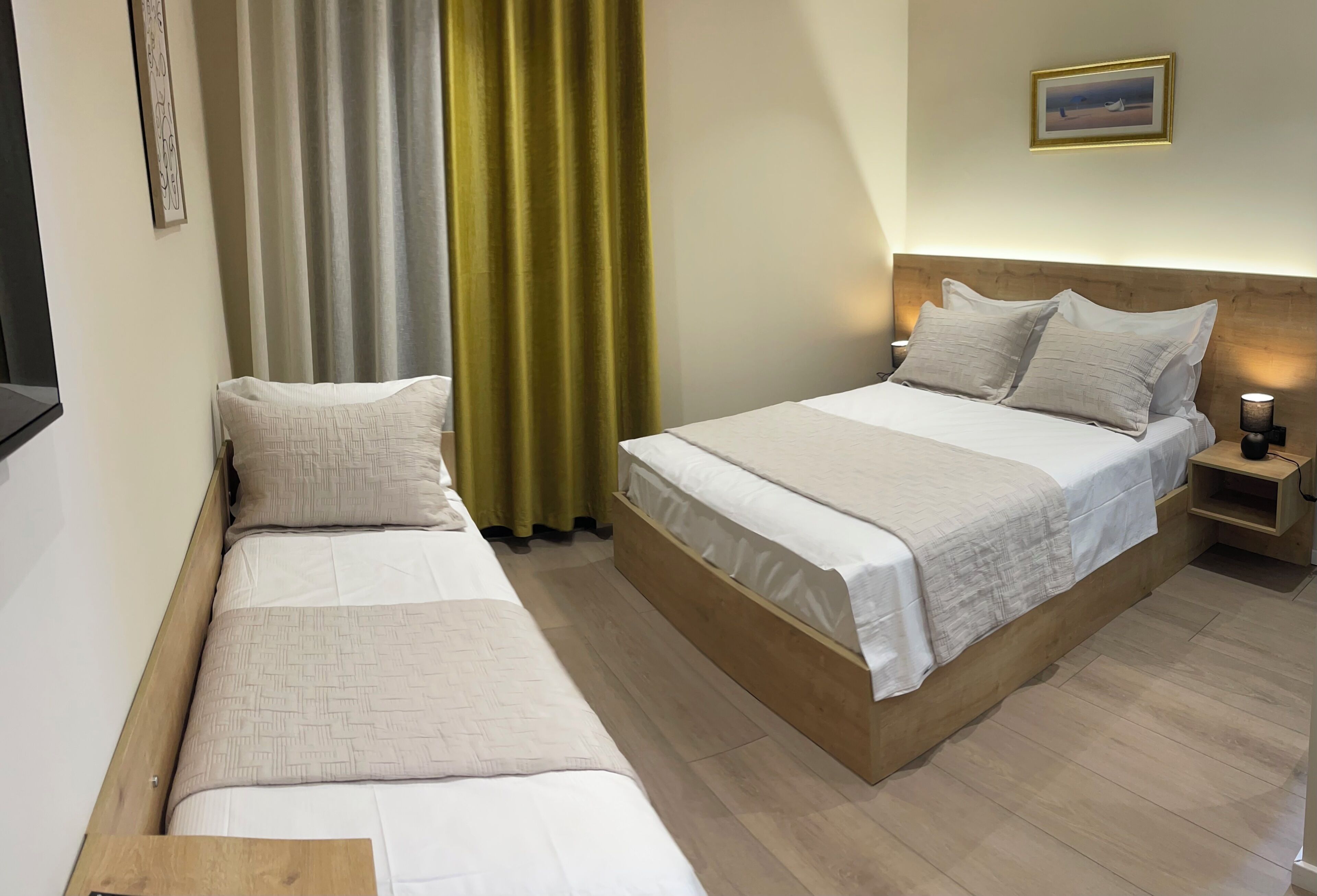 Quarto de hotel com duas camas, cabeceira de madeira, mesa de cabeceira com abajur e um quadro na parede.
