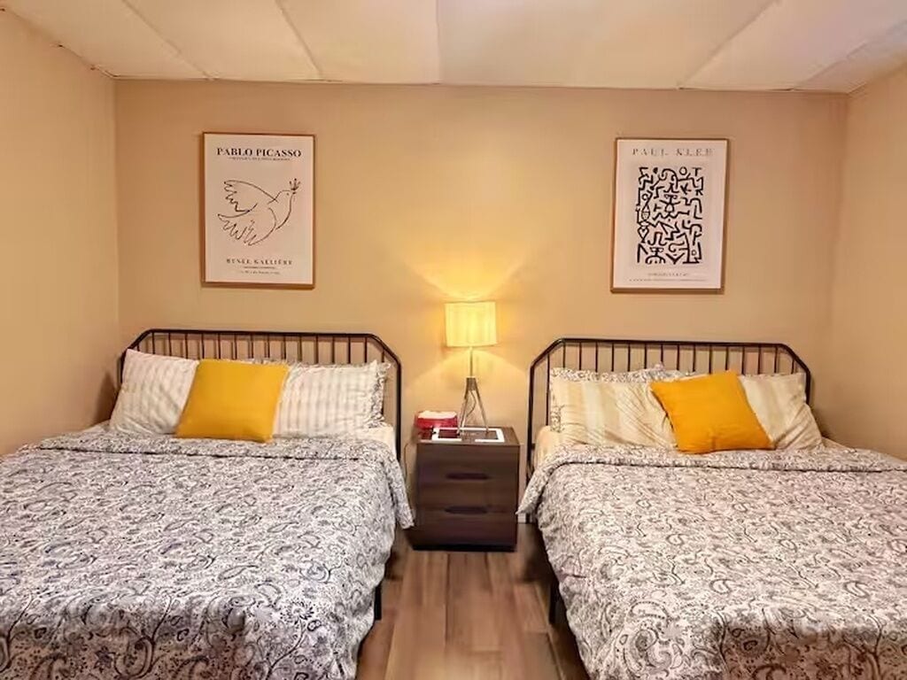 Ein Zimmer mit zwei Betten, jeweils mit gemusterten Bettbezügen und einem gelben Kissen. An den Wänden hängen gerahmte Kunstwerke, ein Nachttisch mit einer Lampe und eine kleine Kommode zwischen den Betten.