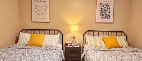 Ein Zimmer mit zwei Betten, jeweils mit gemusterten Bettbezügen und einem gelben Kissen. An den Wänden hängen gerahmte Kunstwerke, ein Nachttisch mit einer Lampe und eine kleine Kommode zwischen den Betten.